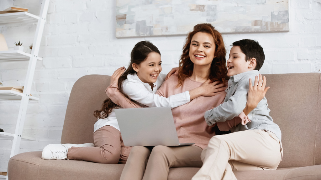 Mosolygó anya ölelés gyerekek közelében laptop a kanapén  - Fotó, kép