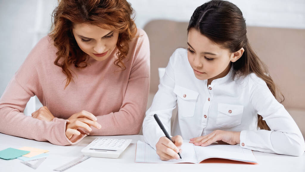 Mädchen schreibt auf Notizbuch neben Mutter und Taschenrechner auf Tisch  - Foto, Bild