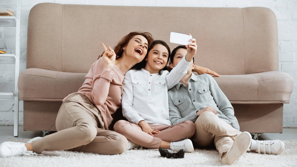KYIV, UKRAINE -  APRIL 15, 2019: Happy family taking selfie on smartphone near joystick on floor  - Foto, afbeelding
