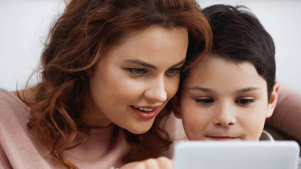 Smiling mother and son using digital tablet on blurred foreground during online education - Photo, Image