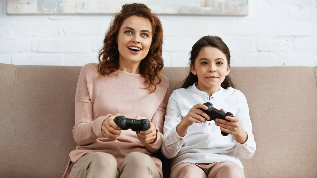 KYIV, UCRANIA - 15 DE ABRIL DE 2019: Madre sonriente jugando videojuegos cerca de la hija preadolescente en el sofá  - Foto, Imagen