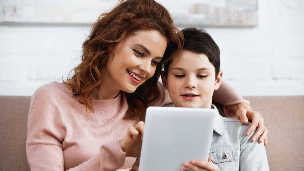 Hymyilevä äiti käyttää digitaalista tablettia sumealla etualalla lähellä poikaa kotona  - Valokuva, kuva