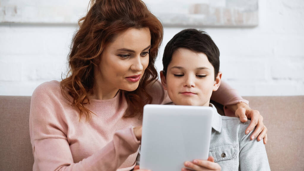 Улыбающаяся женщина обнимает сына и использует цифровой планшет во время онлайн-обучения  - Фото, изображение