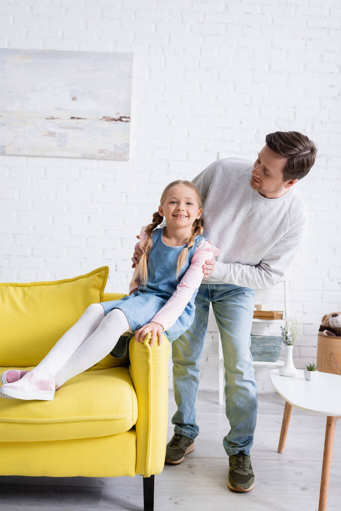 man touching shoulders of cheerful child sitting on sofa in living room - Photo, Image