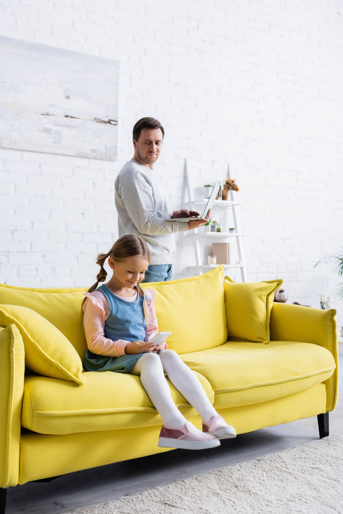 χαμογελαστό κορίτσι κάθεται στον καναπέ με smartphone κοντά στον πατέρα κρατώντας φορητό υπολογιστή - Φωτογραφία, εικόνα