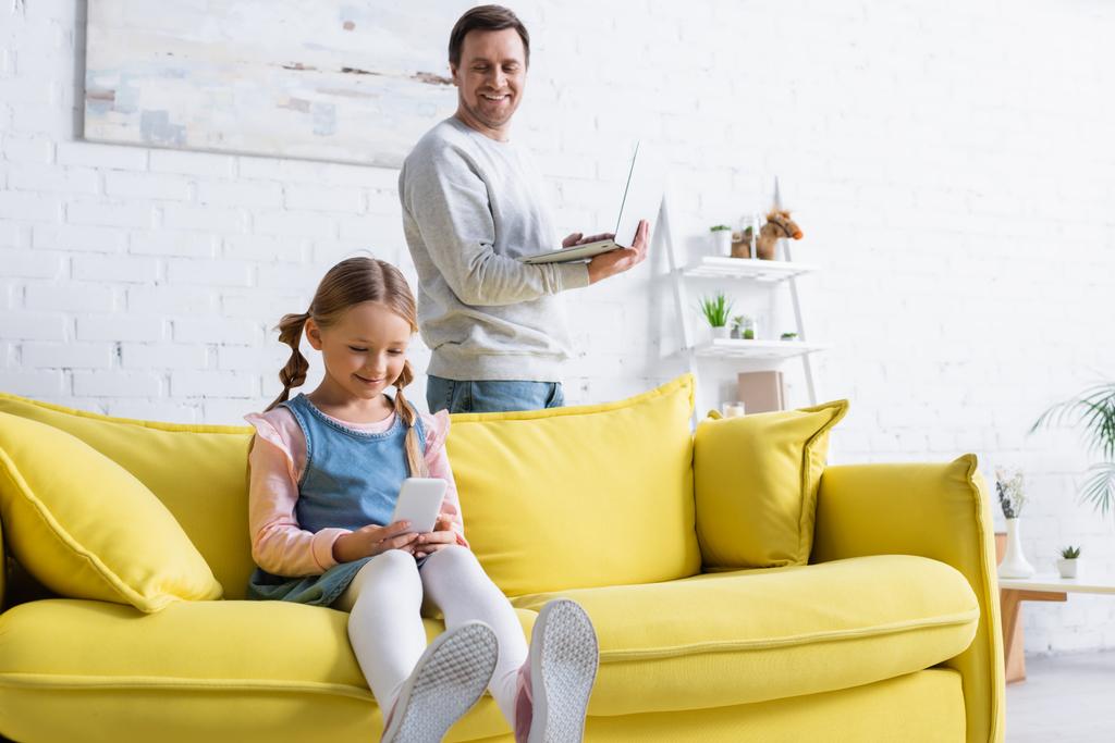 uśmiechnięty mężczyzna z laptopem patrząc na córkę za pomocą telefonu komórkowego na kanapie - Zdjęcie, obraz