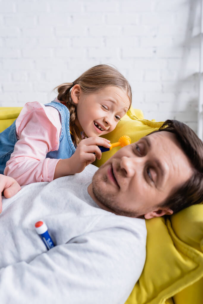 muž ležící na pohovce poblíž dcery zkoumá ho s otoskopem, zatímco si hraje na doktora, rozmazané popředí - Fotografie, Obrázek