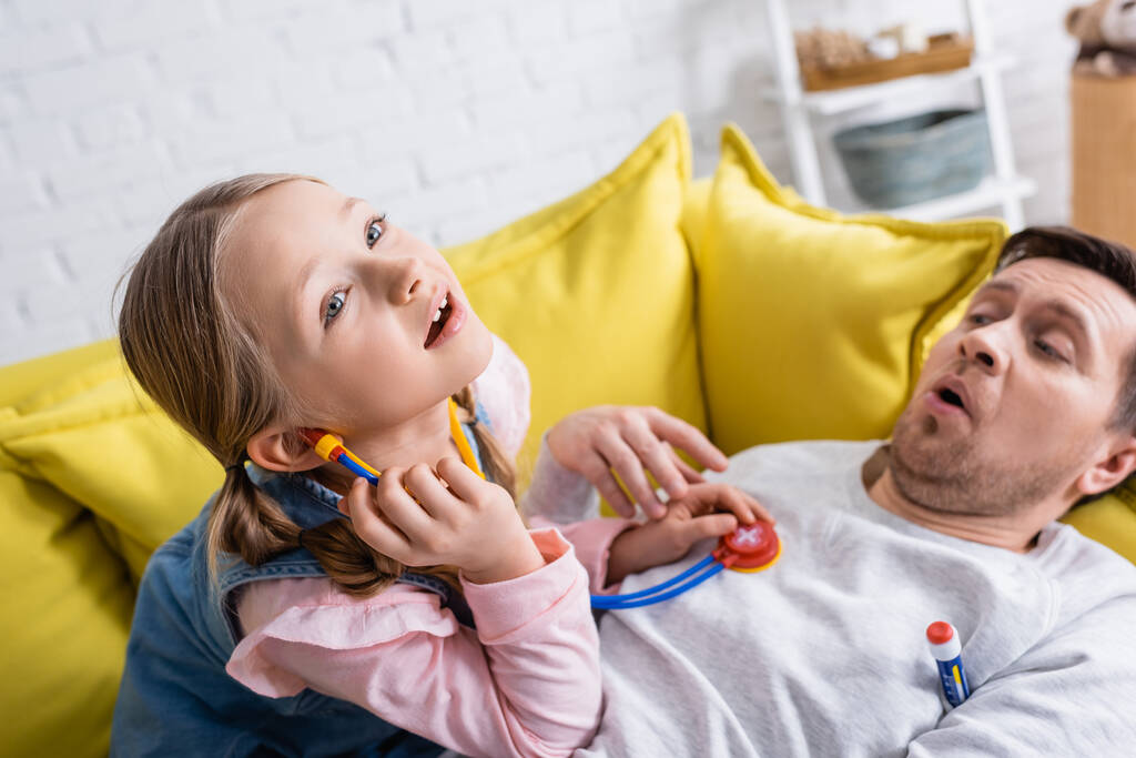 hombre acostado en el sofá y fingiendo estar enfermo mientras su hija lo examina con estetoscopio de juguete - Foto, Imagen