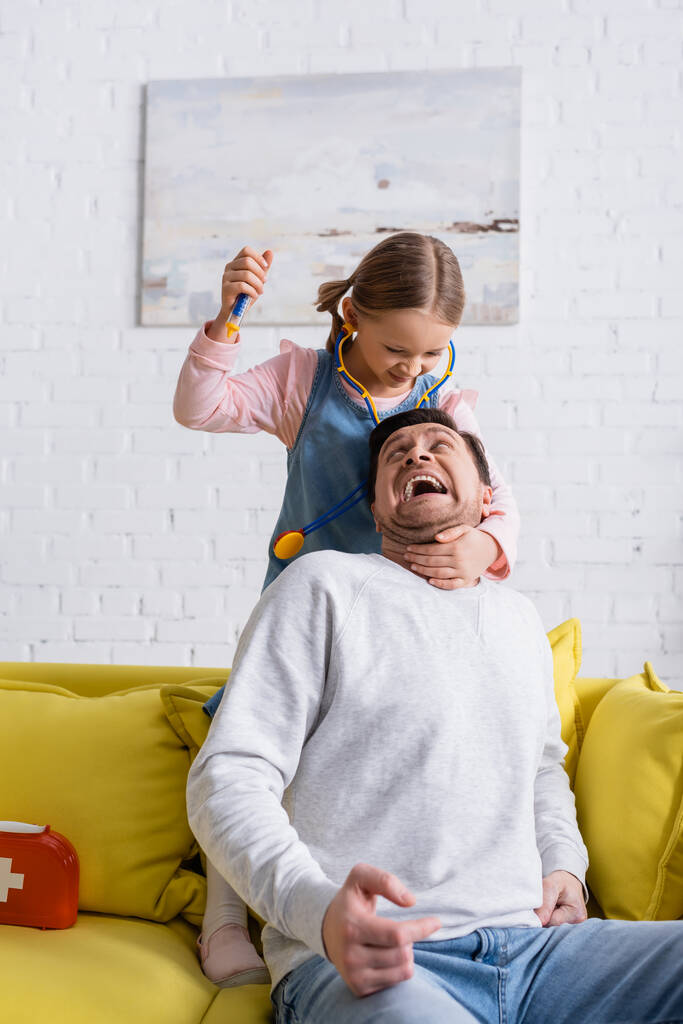Мужчина кричит, притворяясь испуганным рядом с дочерью, держащей игрушечный шприц - Фото, изображение