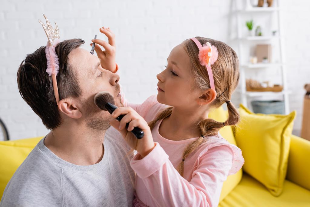 dívka v koruně hračky dělat make-up s kosmetické štětce na otce - Fotografie, Obrázek