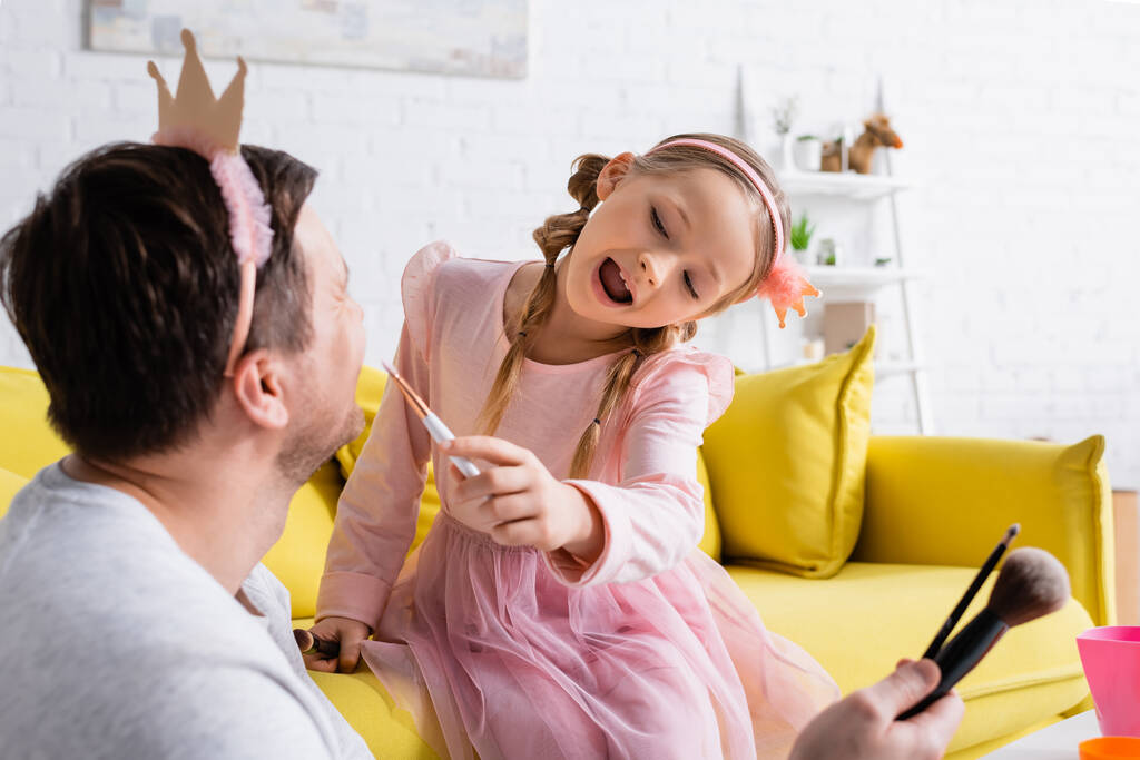 hombre en juguete corona celebración de cepillos cosméticos mientras hija excitada aplicación de maquillaje, primer plano borrosa - Foto, Imagen