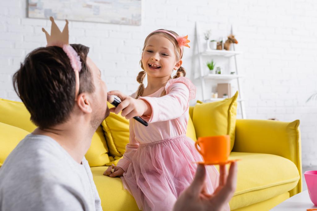 щаслива дівчина в іграшковій короні робить макіяж батькові на розмитому фоні
 - Фото, зображення