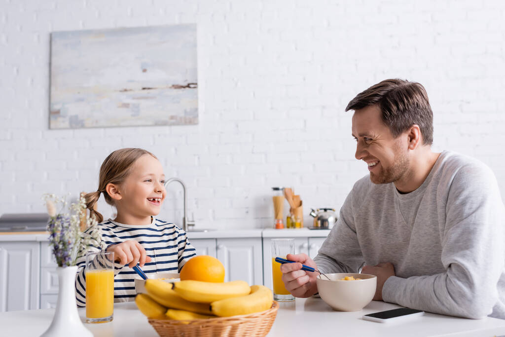 padre e hija sonriendo el uno al otro mientras comen hojuelas de maíz cerca de frutas frescas en primer plano borroso - Foto, Imagen