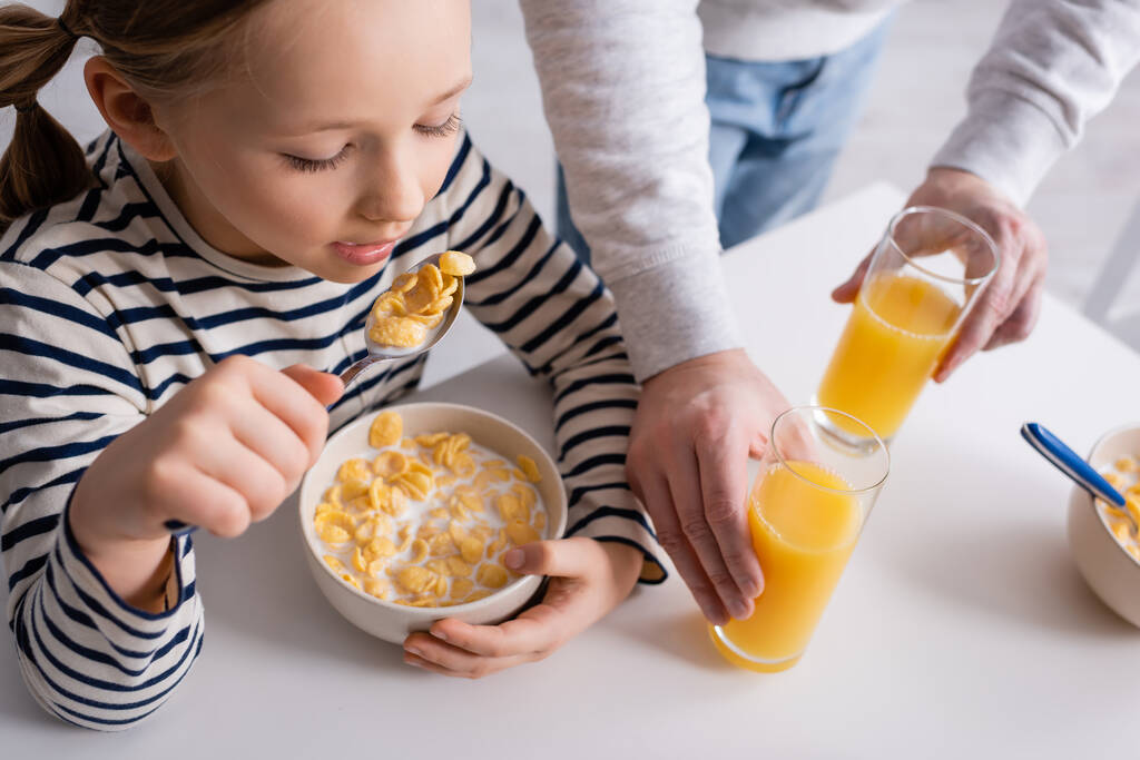 chica comiendo copos de maíz cerca de padre y vasos de jugo de naranja - Foto, imagen