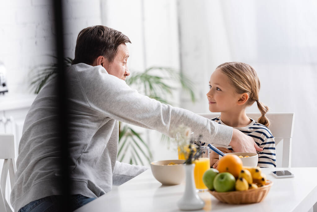 усміхнена дівчина дивиться на батька під час сніданку на кухні, розмитий передній план
 - Фото, зображення
