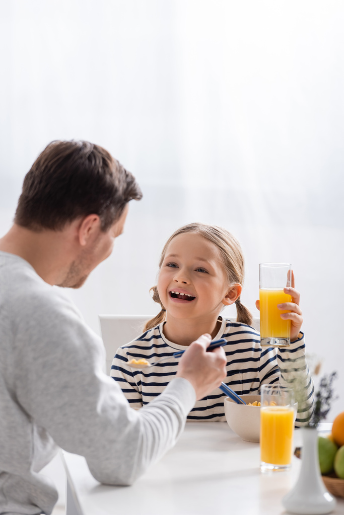 podekscytowane dziecko trzymające szklankę soku pomarańczowego w pobliżu ojca na niewyraźnym pierwszym planie - Zdjęcie, obraz