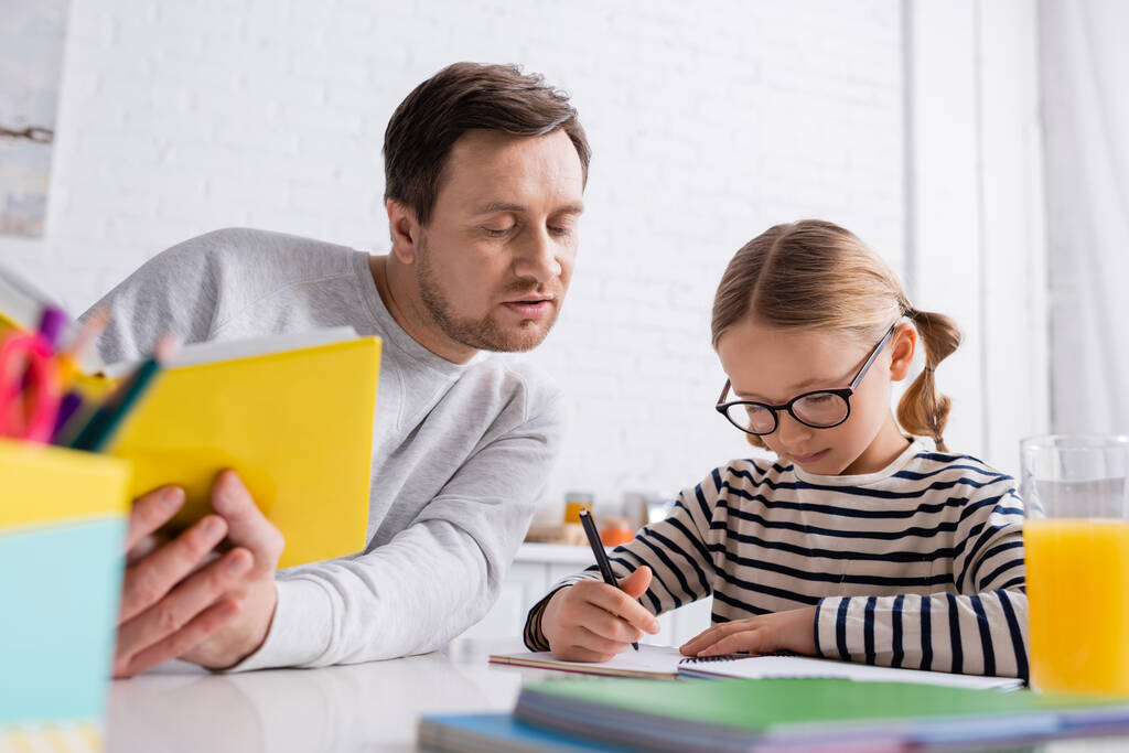 Отец держит книгу рядом с дочерью, записывая в блокнот во время выполнения домашних заданий, размытый передний план - Фото, изображение