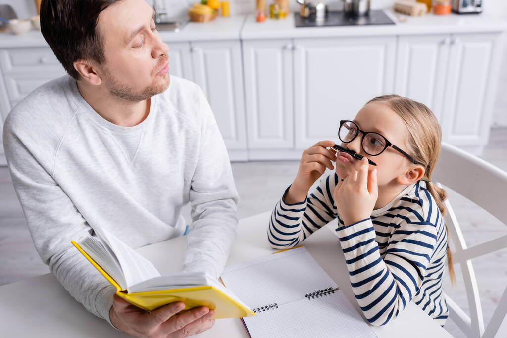 speels meisje houden pen in de buurt van neus terwijl het doen van huiswerk met vader - Foto, afbeelding