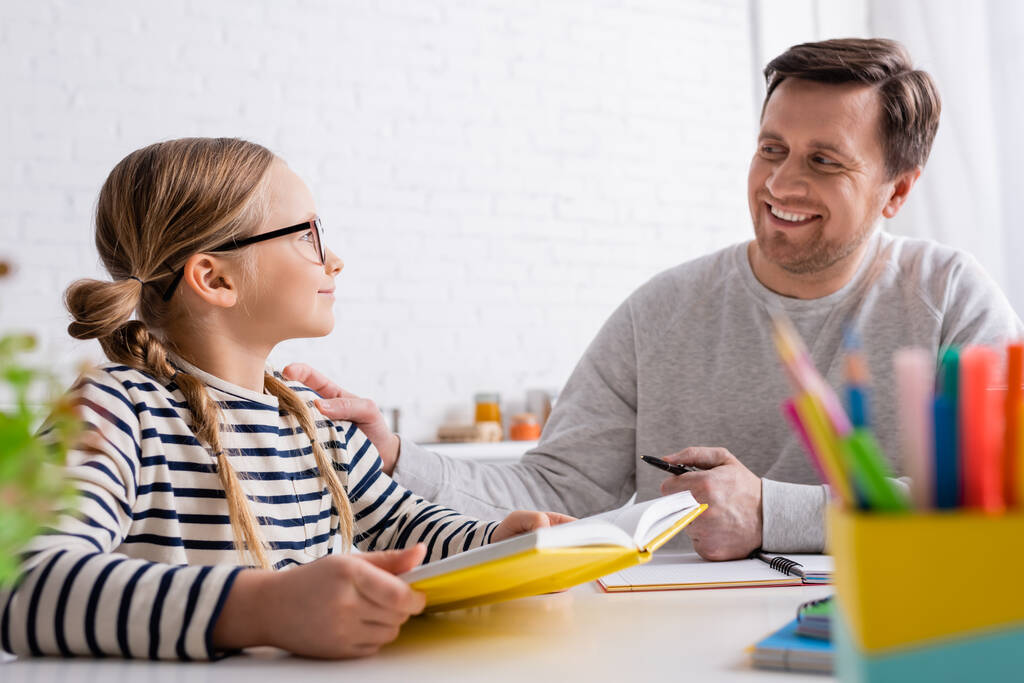 sonriente hombre mirando a su hija sosteniendo el libro de texto mientras hace la tarea en primer plano borroso - Foto, Imagen