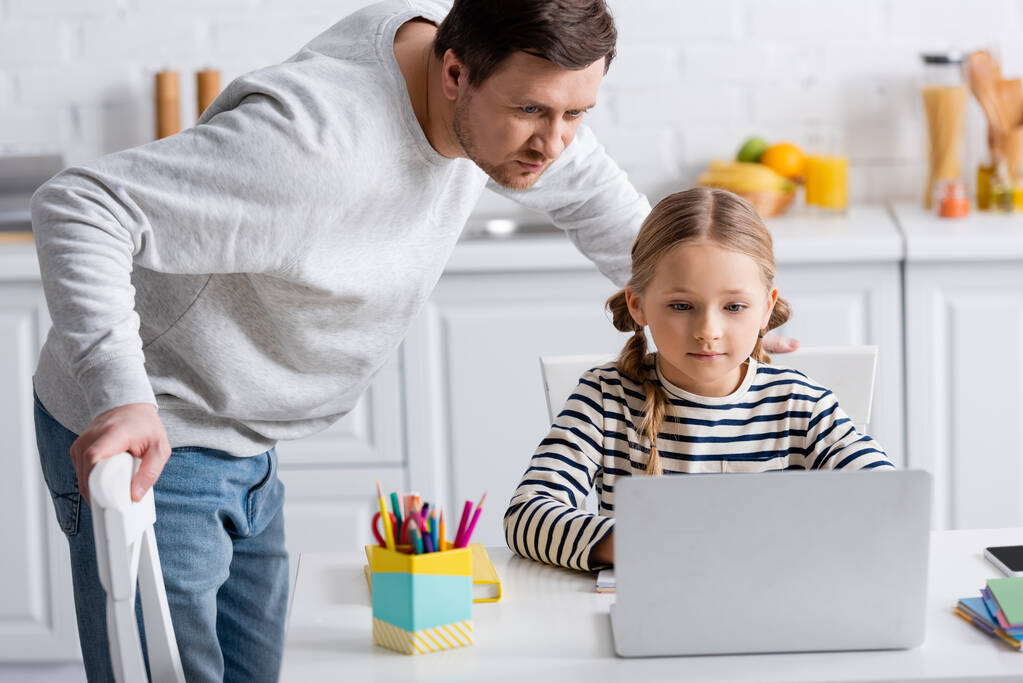girl doing homework near laptop and father in kitchen - Photo, Image