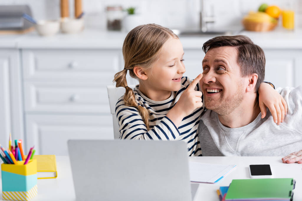 menina alegre tocando nariz do pai enquanto se divertindo perto de laptop na cozinha - Foto, Imagem