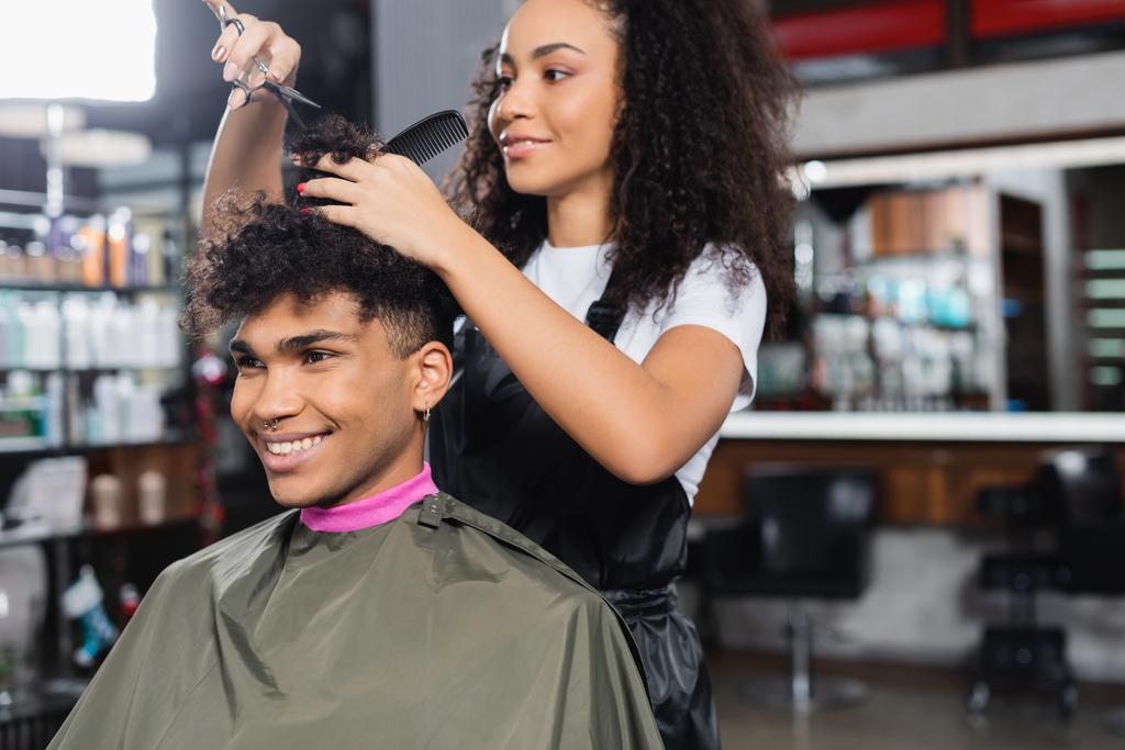 Allegro cliente afro-americano seduto vicino al parrucchiere con forbici e pettine in salone  - Foto, immagini