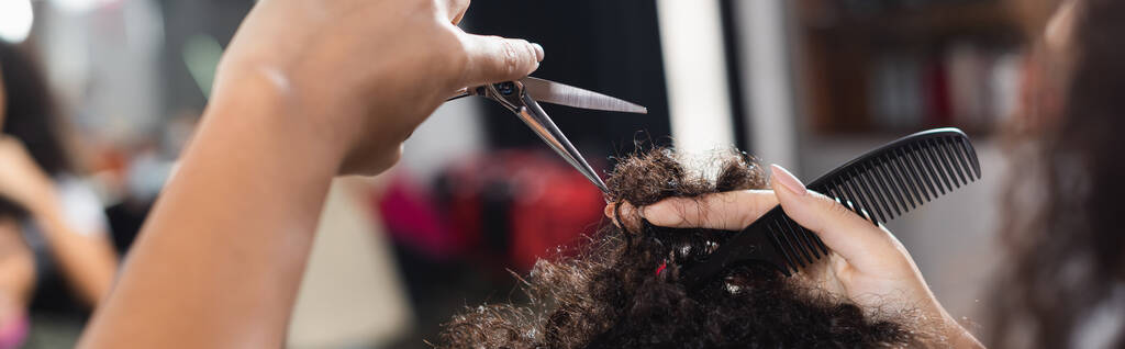 Καλλιεργημένη άποψη του Αφροαμερικανού κομμωτή κρατώντας χτένα και ψαλίδι κοντά στα μαλλιά του πελάτη, πανό  - Φωτογραφία, εικόνα