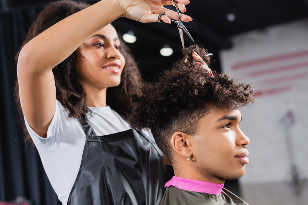 Ciseaux en main de coiffeur afro-américain au premier plan flou travaillant avec le client  - Photo, image