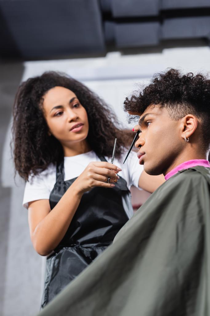 Vue à angle bas du client afro-américain en cape assis près du coiffeur avec des ciseaux sur fond flou  - Photo, image