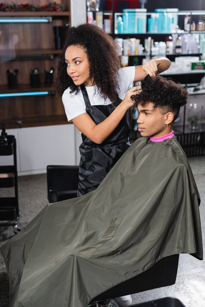 Smiling african american hairstylist combing hair of young man on chair in salon  - Photo, Image