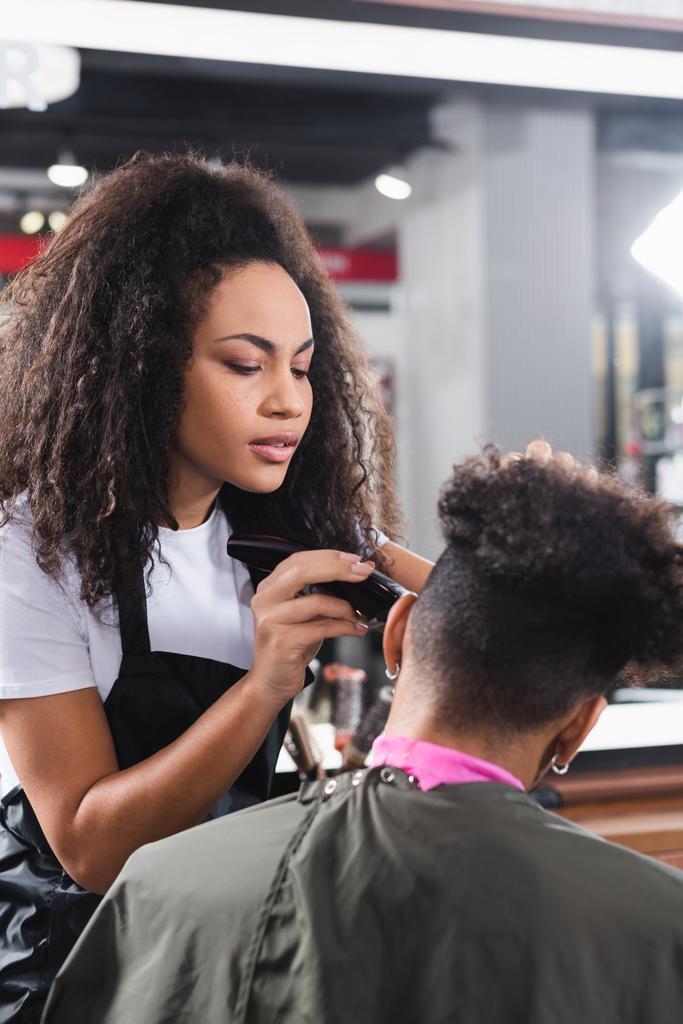 Εστιασμένη Αφροαμερικανός κομμωτής κουρεύει τα μαλλιά του πελάτη στο ακρωτήριο σε θολή πρώτο πλάνο  - Φωτογραφία, εικόνα