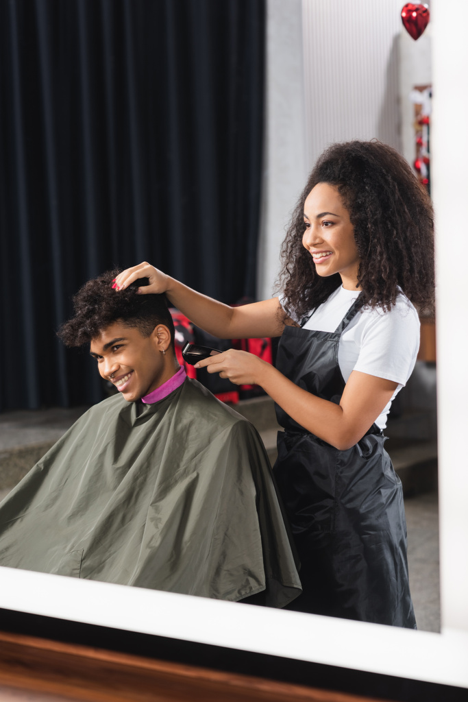Alegre afroamericano hombre sentado cerca de peluquero en delantal celebración trimmer  - Foto, imagen