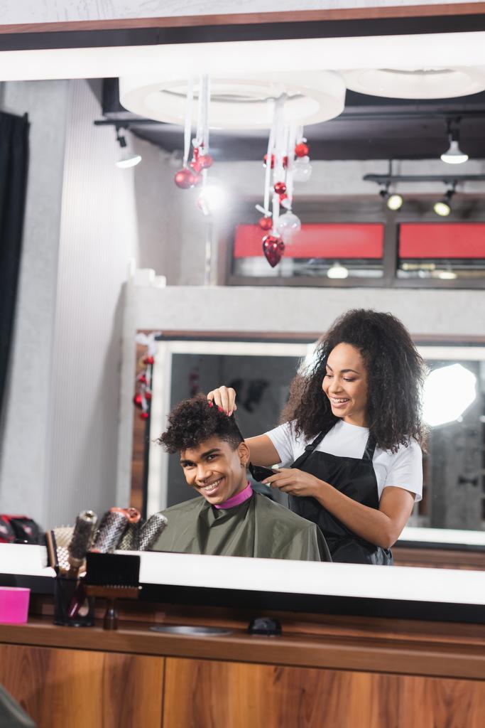 Sonriente afroamericano hombre mirando el espejo cerca de peluquería con trimmer  - Foto, Imagen