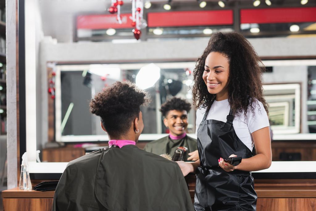 Cliente afroamericano sentado cerca de peluquero sonriente con trimmer en salón  - Foto, imagen