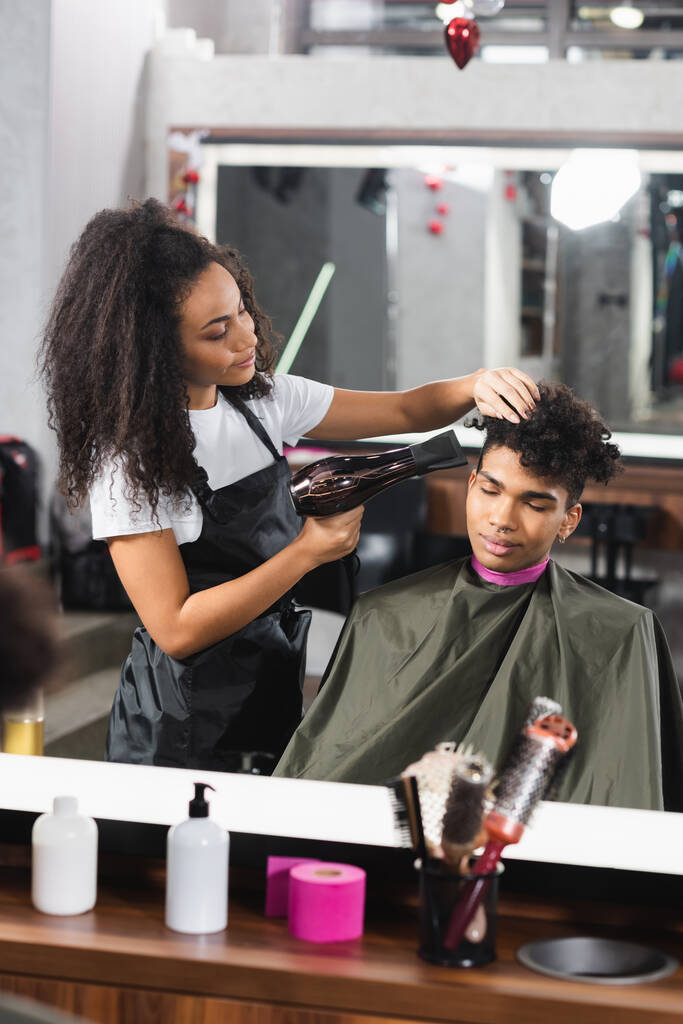 Peluquería afroamericana usando secador de pelo mientras trabaja con el cliente  - Foto, Imagen