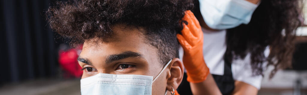 背景のぼやけた医療マスクで美容師の近くに座っているアフリカ系アメリカ人の男性の作物のビュー、バナー  - 写真・画像