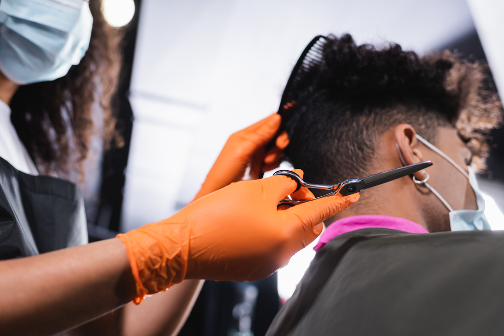 Blick auf die Schere in der Hand eines afrikanisch-amerikanischen Friseurs in der Nähe einer verschwommenen Kundin in medizinischer Maske  - Foto, Bild