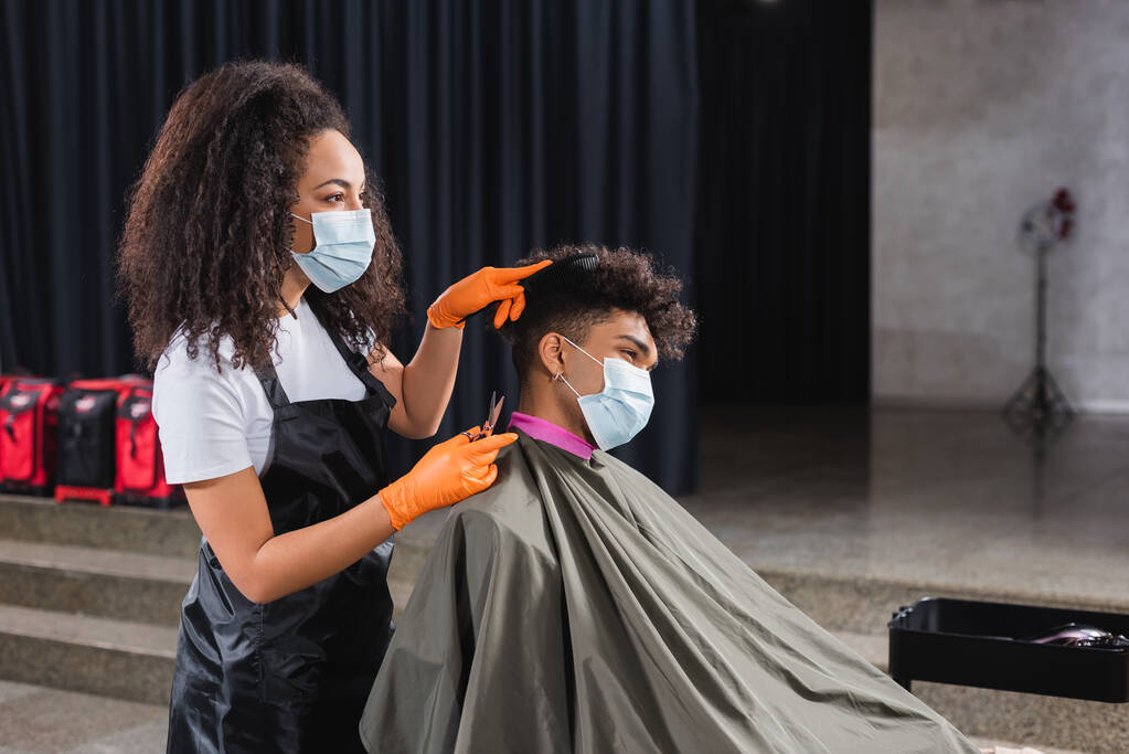 Coiffeur afro-américain en masque médical et gants en latex tenant des ciseaux et peignant les cheveux du client  - Photo, image