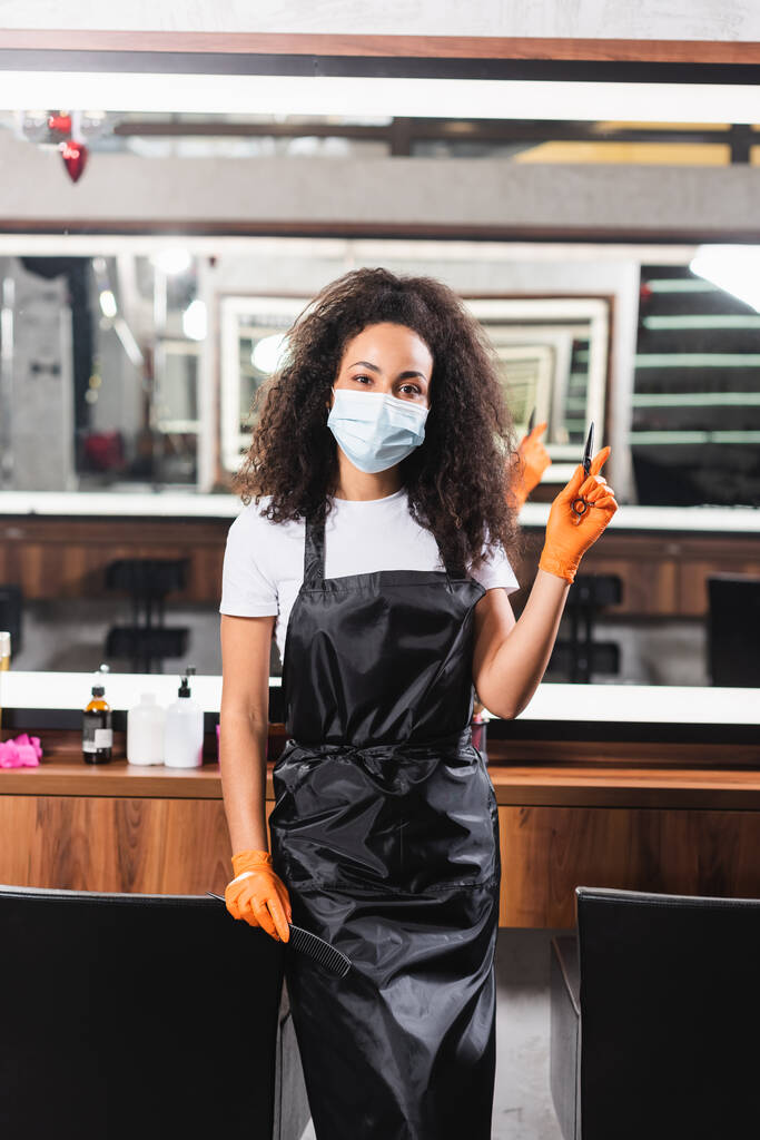 Afrykański amerykański fryzjer w masce medycznej trzymając nożyczki i grzebień wskazując palcem w salonie  - Zdjęcie, obraz