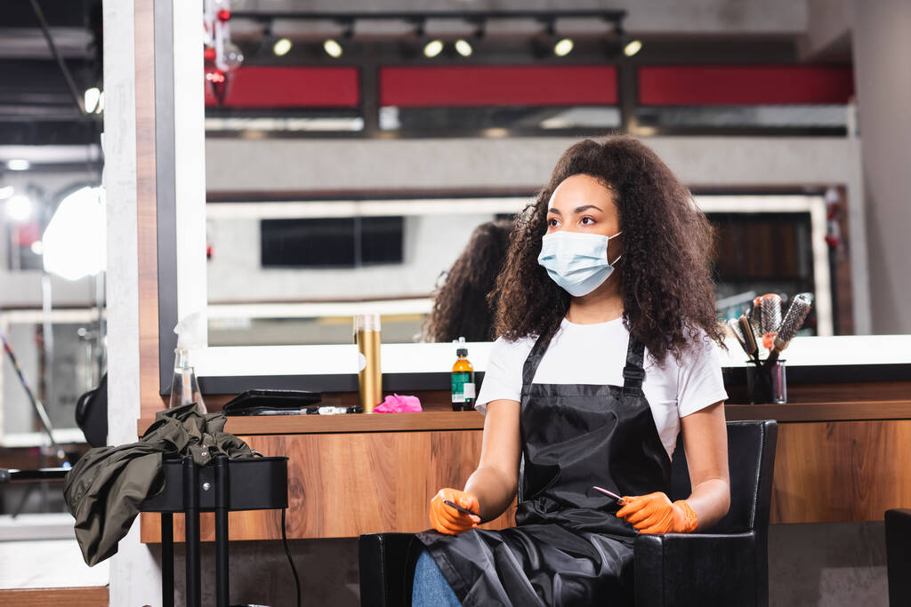 Cabeleireiro afro-americano em máscara médica e luvas de látex segurando tesoura e pente  - Foto, Imagem