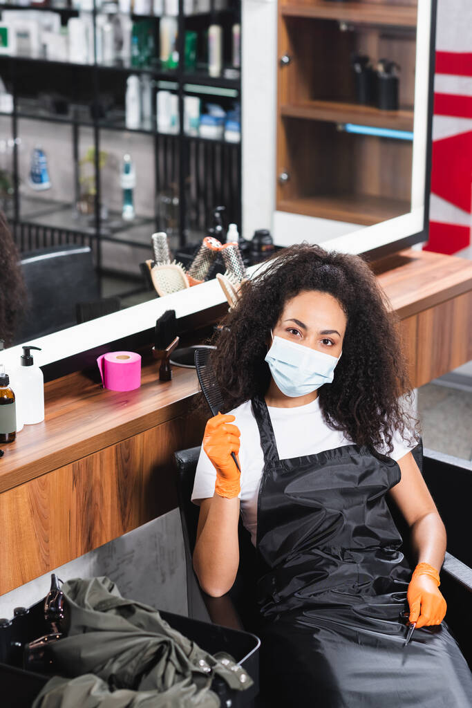 Африканский американский парикмахер в защитной маске и перчатках держит ножницы и расческу возле зеркала в салоне  - Фото, изображение