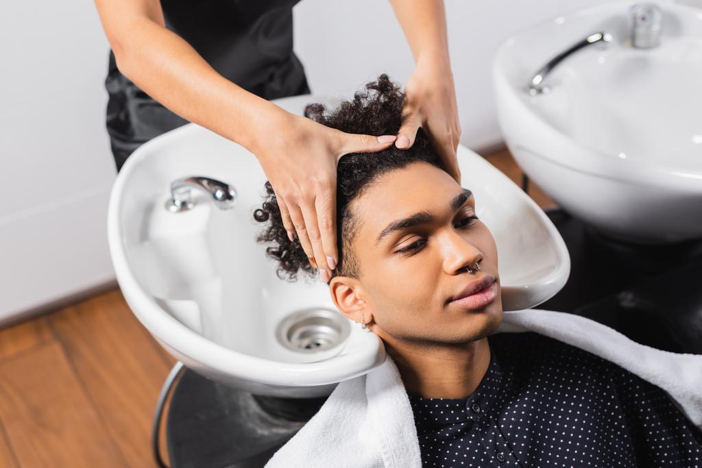 Afroamerikaner sitzt neben Friseur und wäscht Haare und Waschbecken auf verschwommenem Hintergrund - Foto, Bild