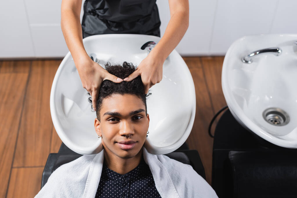 Vue grand angle de l'homme afro-américain regardant la caméra près du coiffeur et l'évier flou dans le salon  - Photo, image