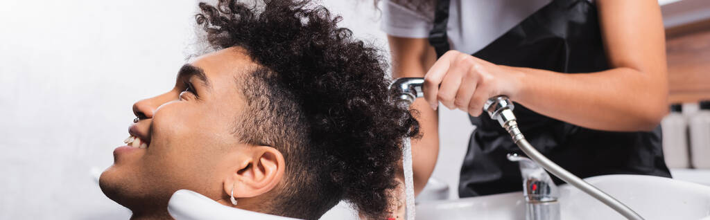 Sonriente cliente afroamericano sentado cerca de peluquería con ducha sobre fondo borroso, pancarta  - Foto, imagen