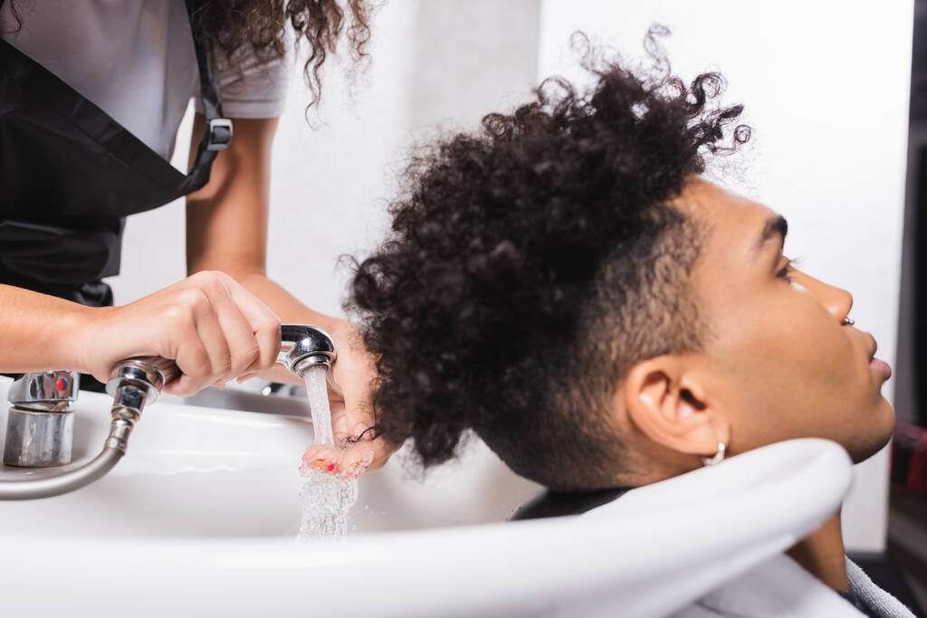 サロンでアフリカ系アメリカ人の美容師と若いクライアントの手でシャワー  - 写真・画像