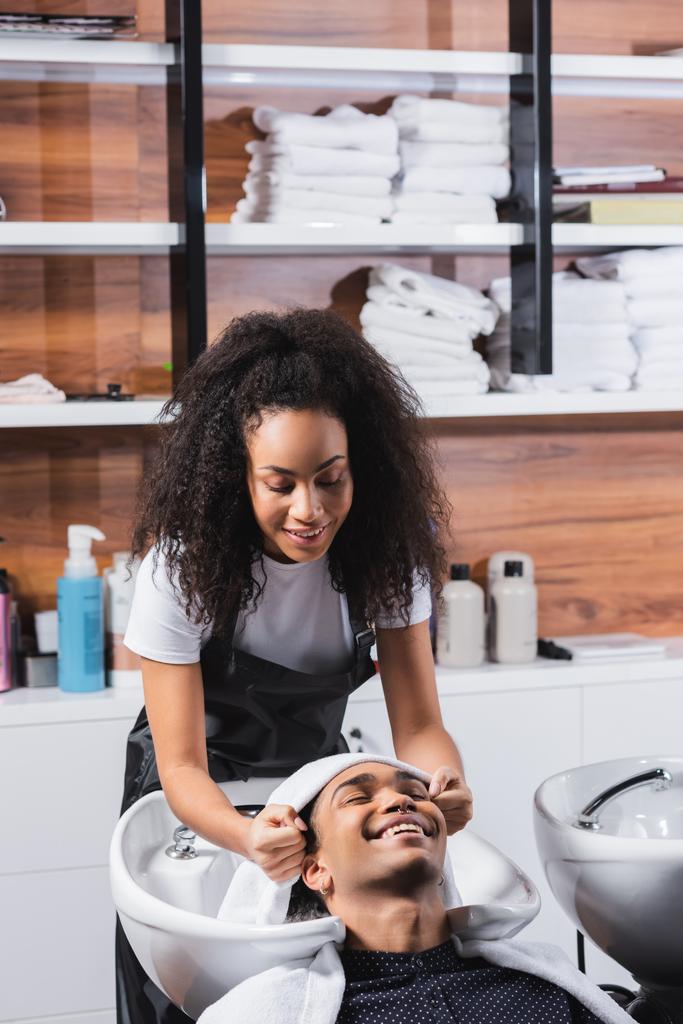 Улыбающийся африканский американец сидит рядом с парикмахером с полотенцем и раковиной  - Фото, изображение
