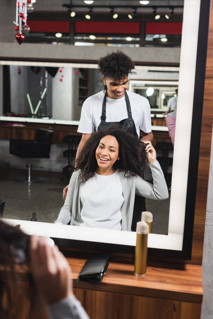 Smiling african american woman touching hair near hairstylist in salon  - Photo, Image