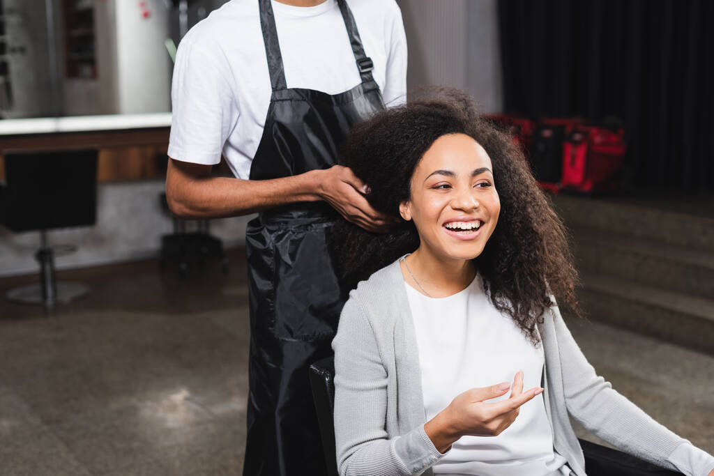 Alegre cliente afroamericano sentado cerca de peluquero en delantal  - Foto, Imagen