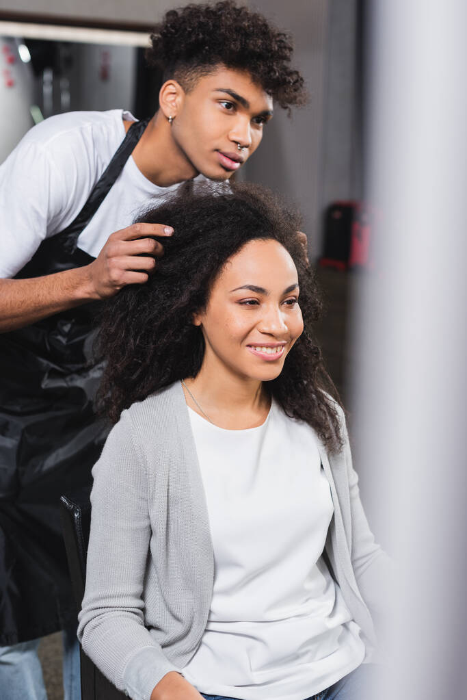 Mujer sonriente sentada cerca de peluquero afroamericano en delantal  - Foto, imagen