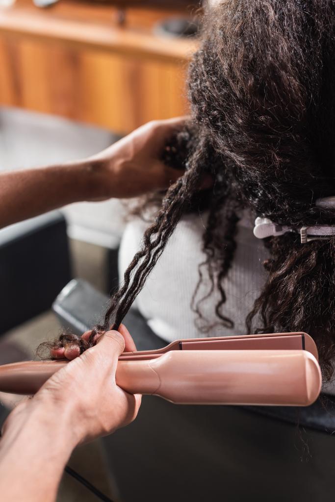 Vista ritagliata del parrucchiere afroamericano che tiene la piastra dei capelli vicino al cliente  - Foto, immagini